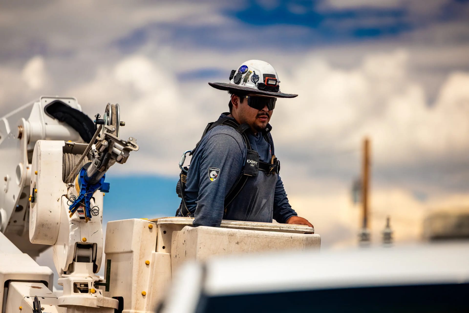 A utility worker in a brimmed hard hat and sunglasses standing in a bucket truck
