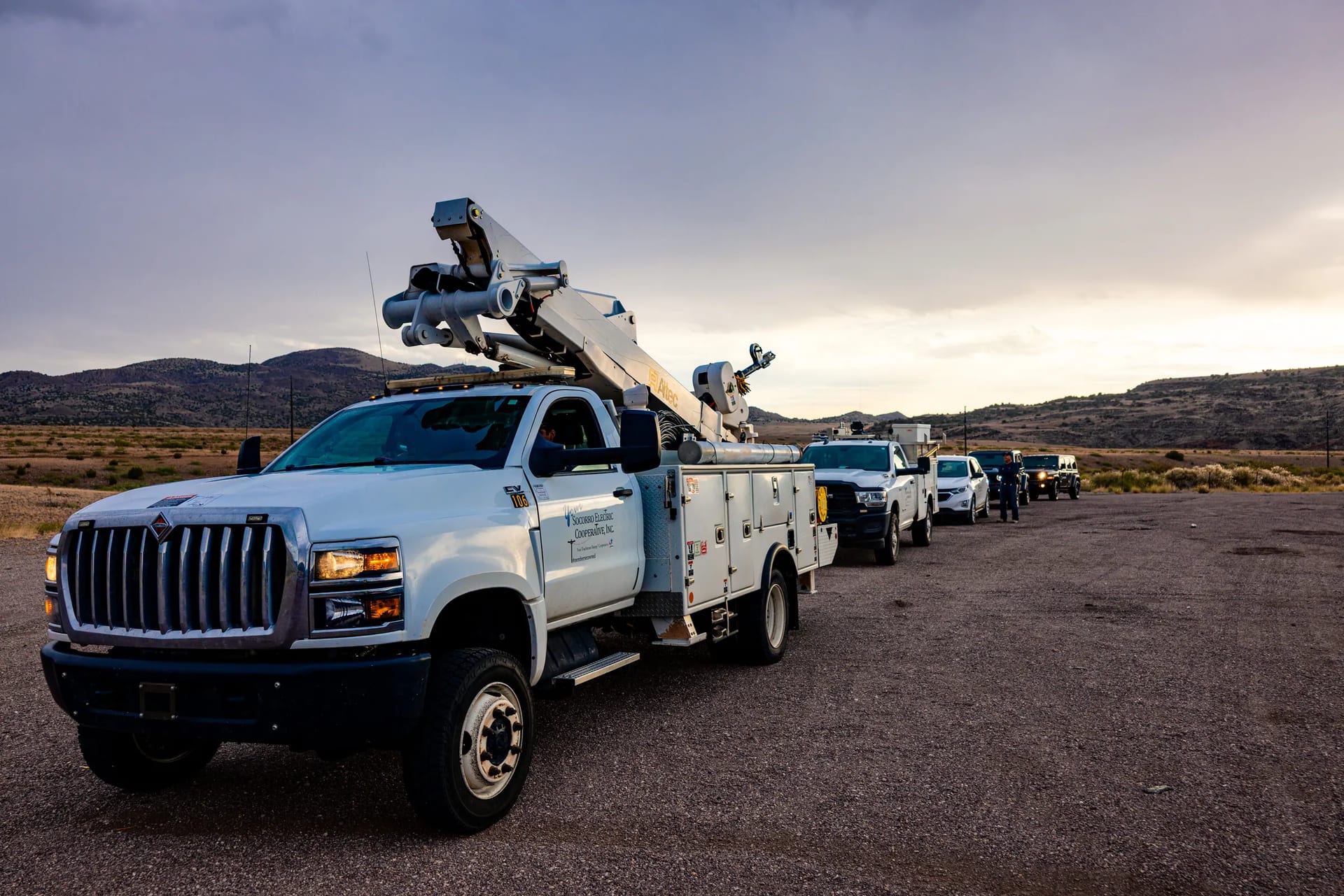 Socorro Electric bucket trucks lined up on a gravel road beneath a cloudy sky